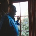 An older man in a blue shirt gazing quietly out of a sunlit window