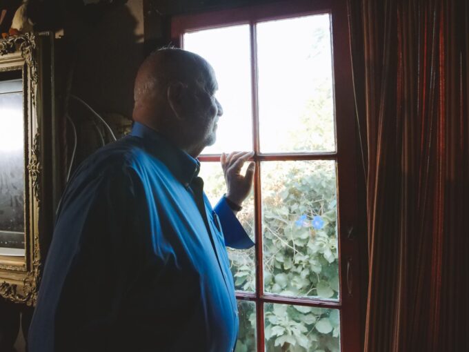 An older man in a blue shirt gazing quietly out of a sunlit window